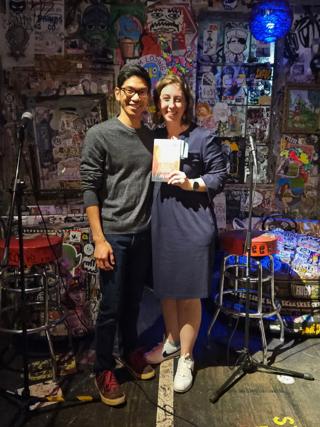 A man and a woman stand side-by-side, the woman holding up a copy of her book.