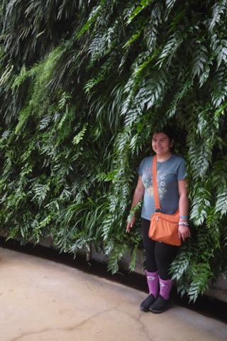 A young woman presses herself into a wall decorated with ferns and leaves in a callback to the Homer hedge meme.
