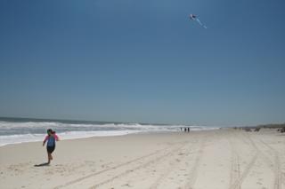 A young woman runs along the beach, trailing a kite behind her".