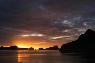 A group of islands silhouetted as the sun sets behind an island to the left.