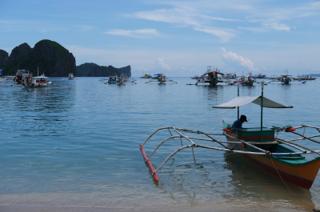 A group of outrigger boats anchored close to shore. In the boat in the foreground a man sits in the shade of a canopy.