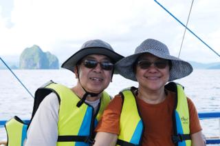 A man and a woman in sunglasses and hats, on a boat, smile with islands in the distance.