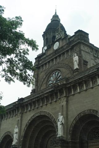 A multi-arched entrance to a stone church, with statues of Catholic saints mounted on pillars to the side of each arch.