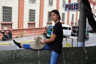 A young girl perches on a wall outside a police station. In her hands she is affixing a single flower to a straw hat.