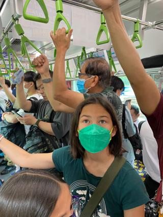 Shot from inside a subway car: a girl wearing a mask looks up at the signs along the car’s roof.