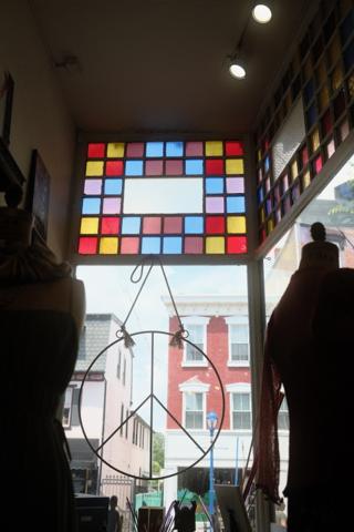 A view from inside a shop window. The upper third of the window is decorated with stained glass in a square pattern. Hanging from a hook is a large peace sign.