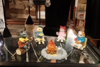 A quartet of cat figurines in human clothing sit in camp chairs, roasting marshmallows around a sculpted fire.