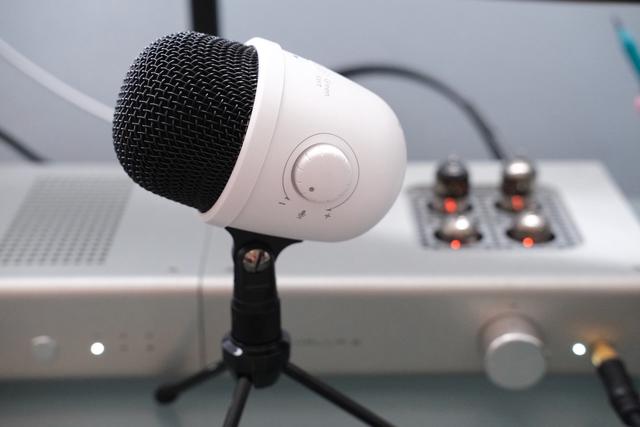 Side view of a white USB microphone, with a headphone amplifier in the background.