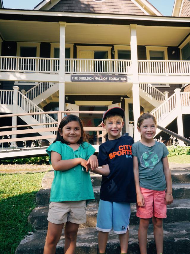 Three young kids pose in front of a school hall.