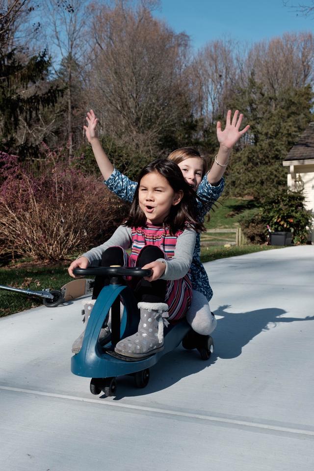 Two girls on a scooter rolling down a driveway.