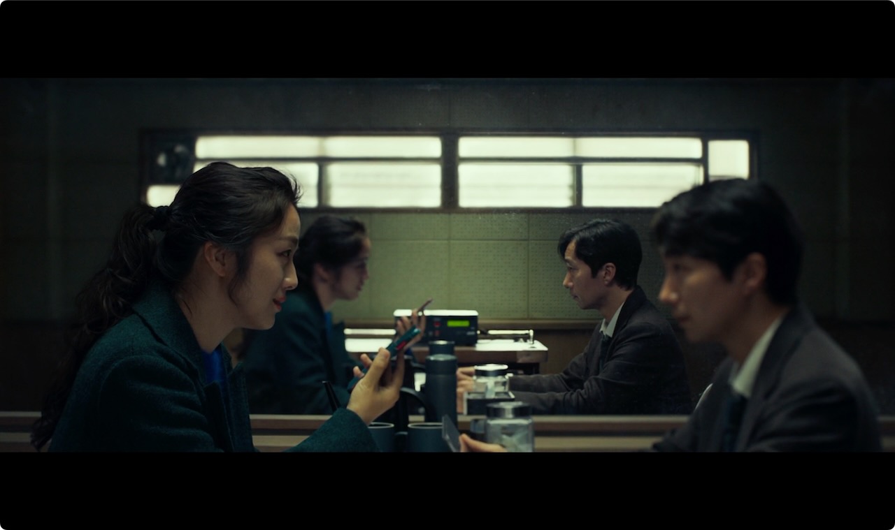 Diagonal split: Seo-rae in foreground focus as she speaks into her translation app. *Image © Mubi* A man and a woman sit facing each other. The woman speaks into a phone.