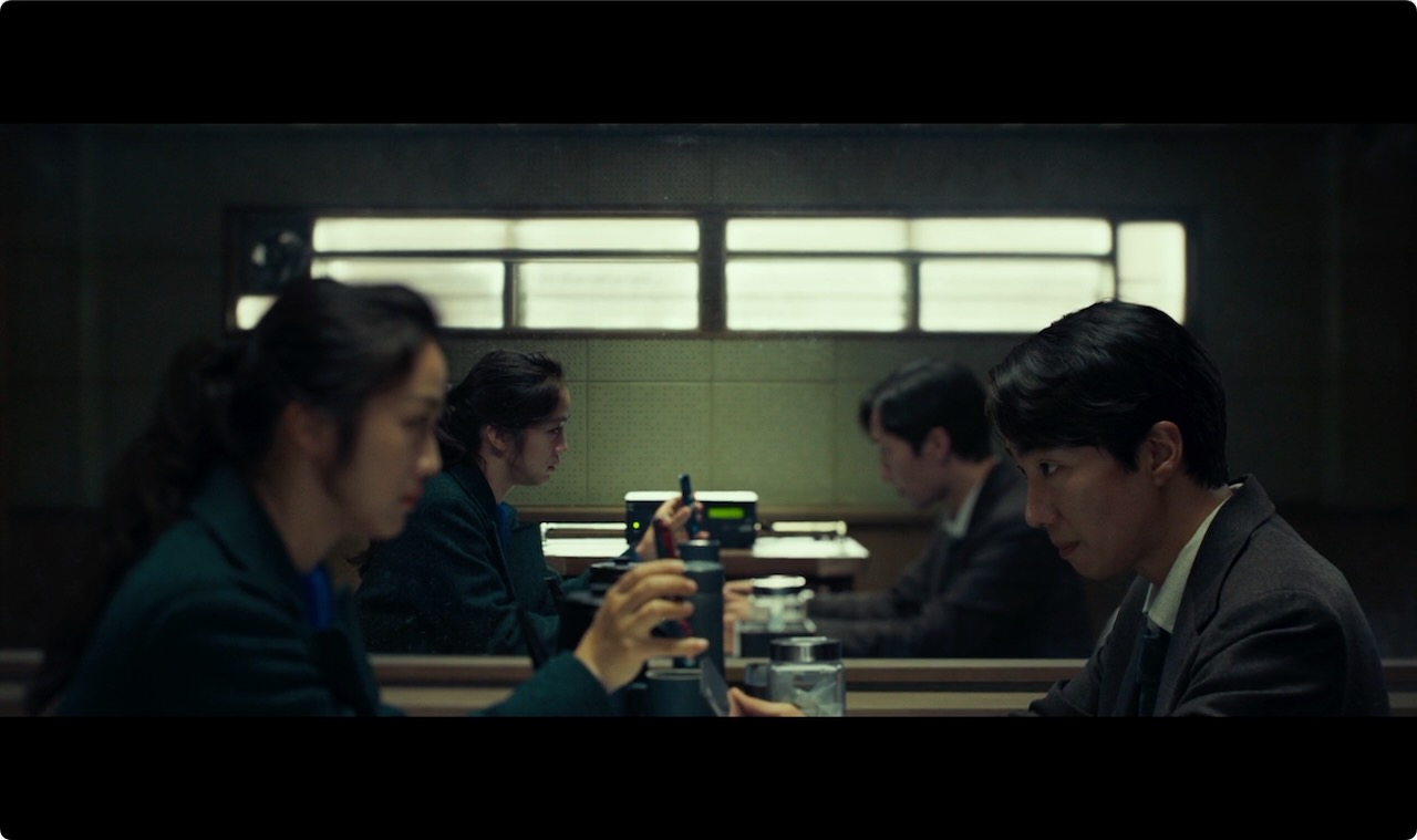 Diagonal split: Seo-rae in background focus as the translation app plays. *Image © Mubi* A man and a woman sit facing each other. The woman holds up her phone between them, playing back a recording.