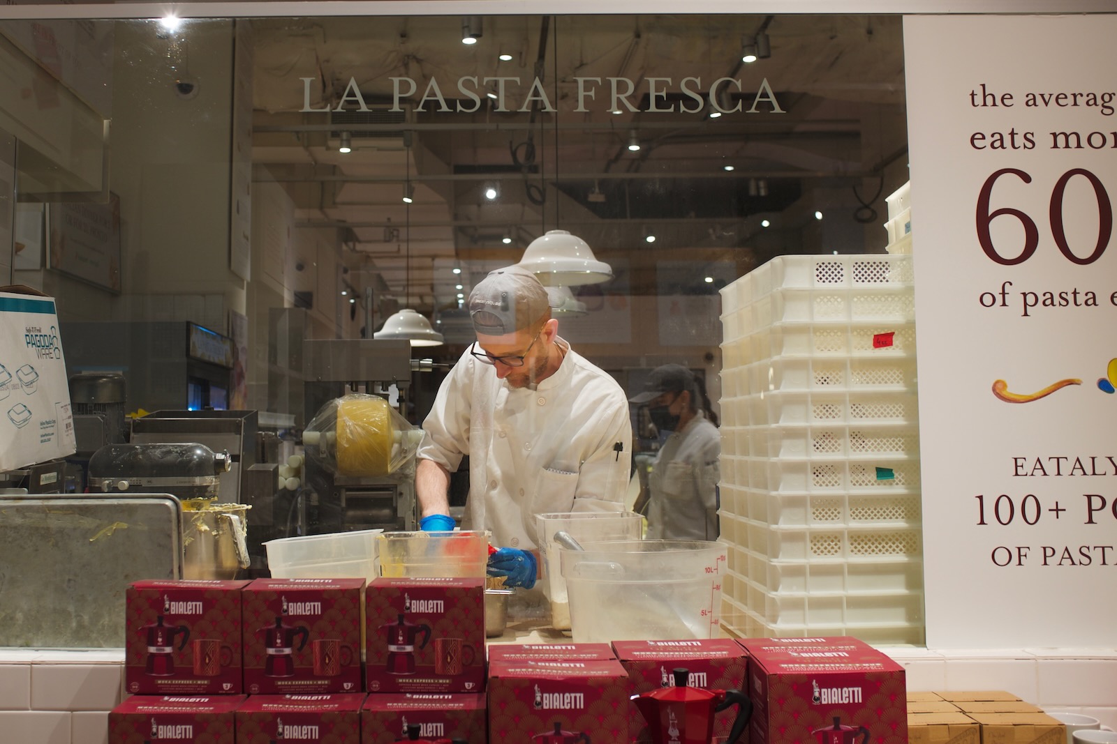 A man in a gray baseball cap and glasses prepares homemade pasta behind a glass partition.