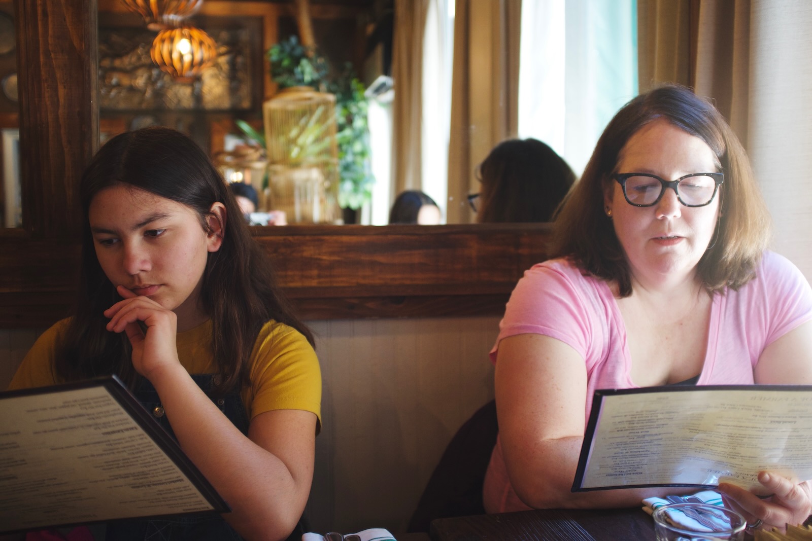 A teenage girl and a woman read menus. Behind them a mounted mirror reflects a restaurant interior.