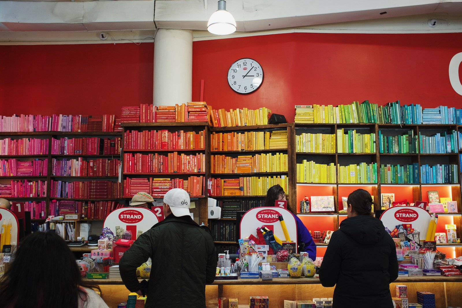 Strand bookstore in New York City. At the checkout counter, a wall of bookshelves decorated with books organized by color extends behind the cashiers.