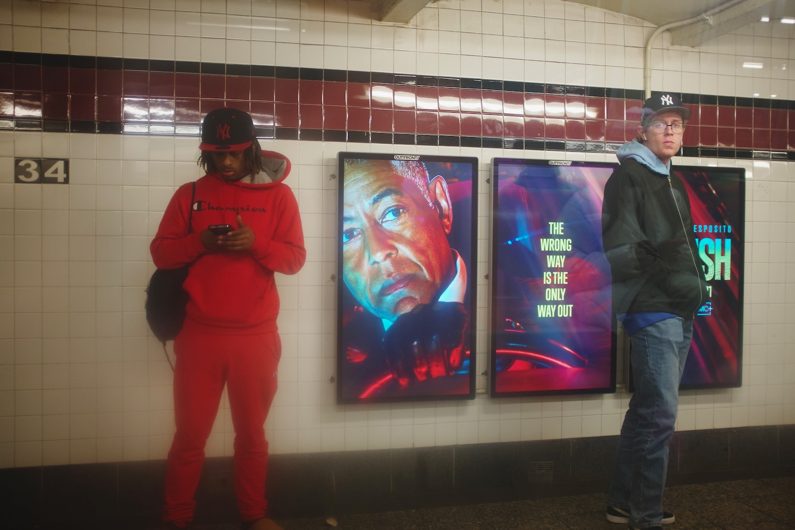 Two men on a subway platform, shot from inside a subway car. The man on the left wears a red hoodie and a black and red Yankees cap. The man on the right wears a traditional blue and white Yankees cap.”