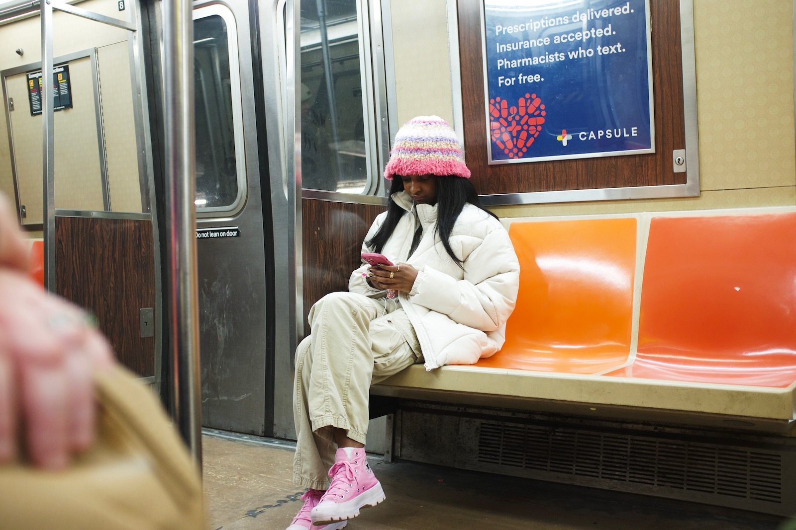 A woman in a fuzzy, multicolored bucket hat and pink shoes looks at her phone while riding the subway.”