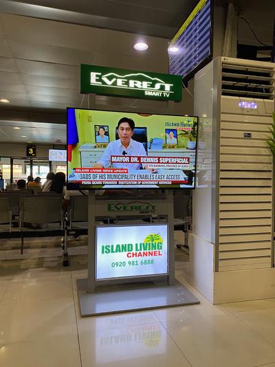 A tv in an airport waiting area. Onscreen a chyron identifies a man onscreen as Mayor Dr. Dennis Superficial