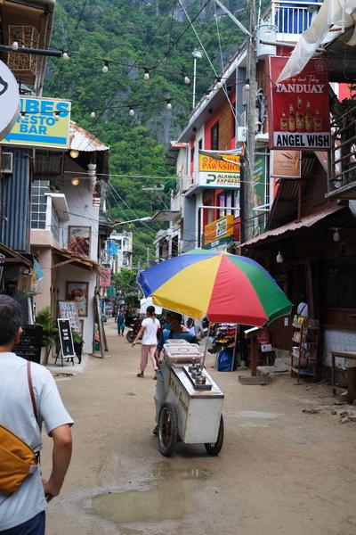 An ice cream cart with a color wheel umbrella stops in the middle of a dirt road lined with shops.