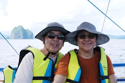 A man and a woman in sunglasses and hats, on a boat, smile with islands in the distance.