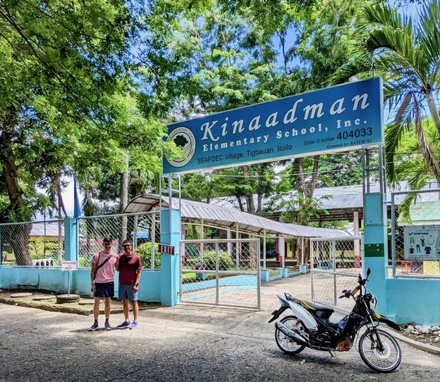 Two men stand in front of a gated entrace to an elementary school called Kinaadman.