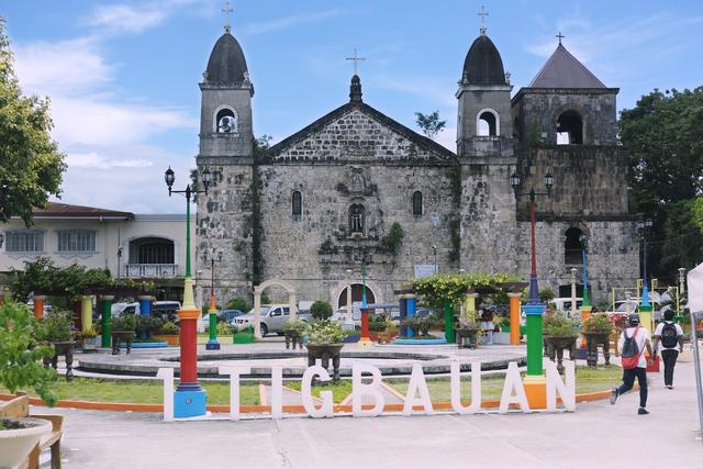 A Catholic church from the 1500s with a sign “Tigbauan” in the plaza in front of it.