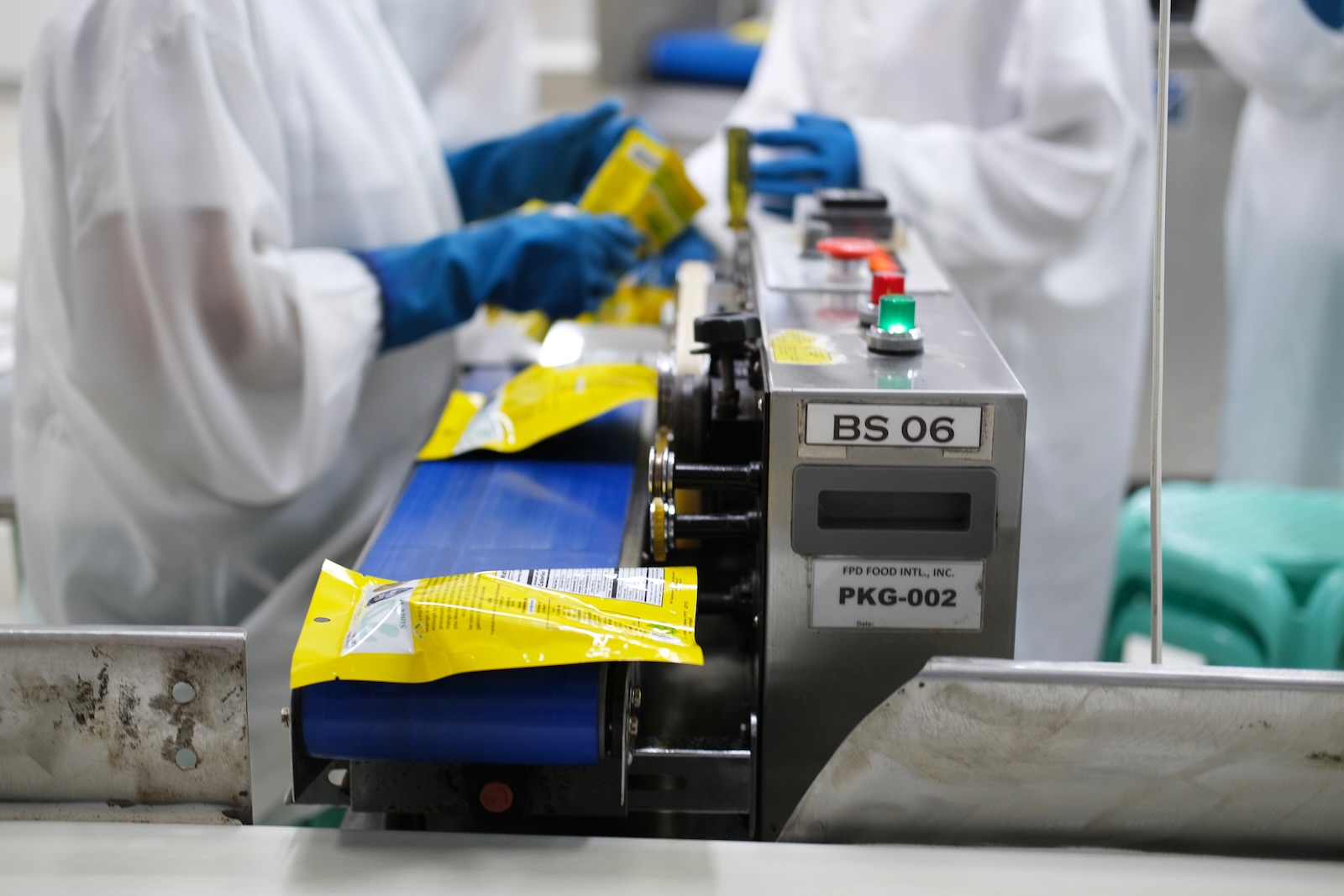 A row of bags containing dried mangoes on a conveyor belt passing through a sealing machine.