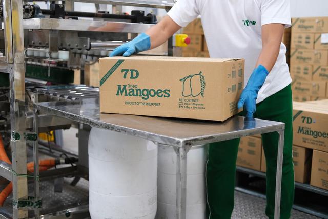 A worker lifts a cardboard box stamped with “7D Mangoes” on its side.