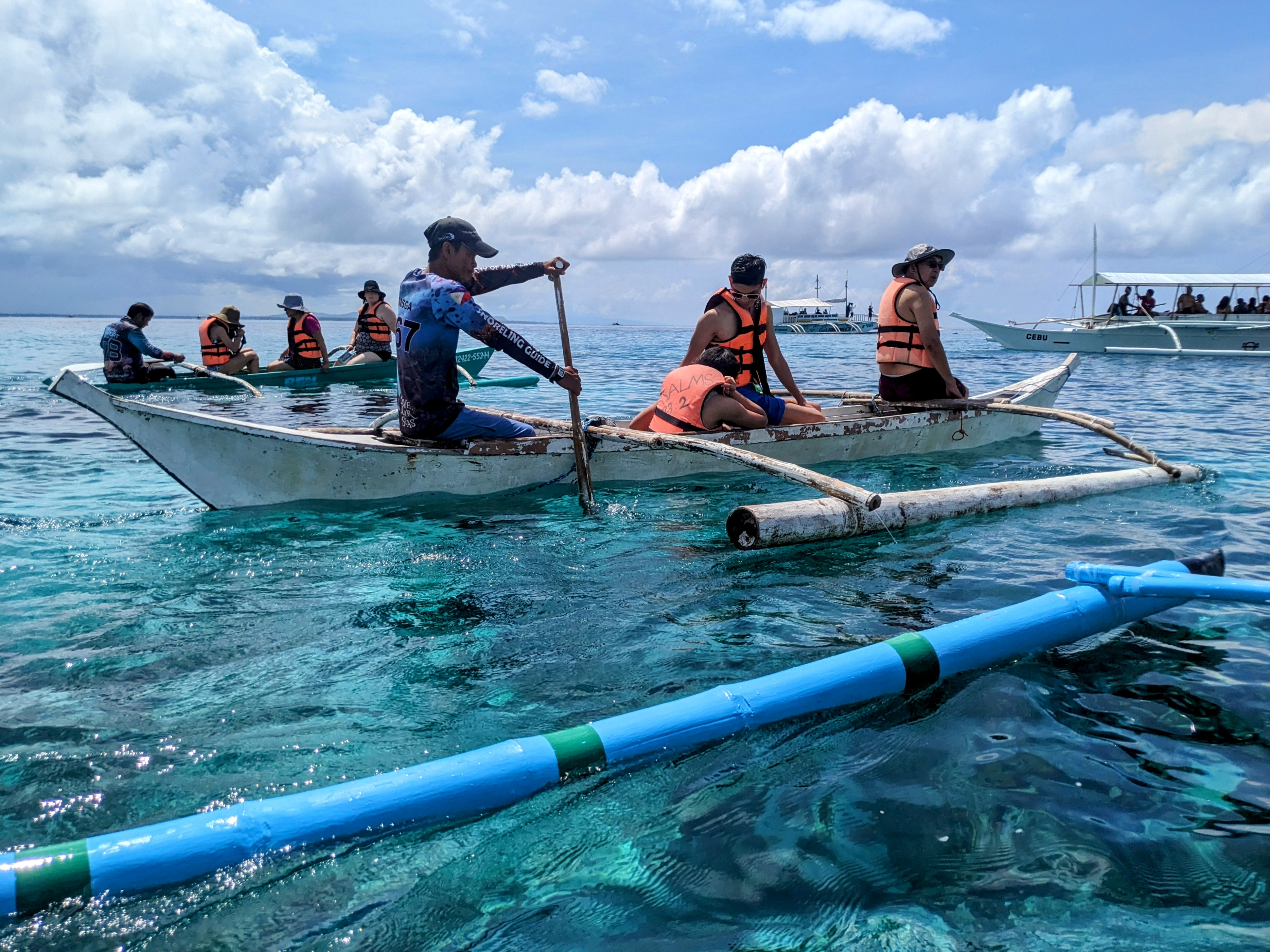 Photo © Joseph Llobrera Looking over the port side of an outrigger boat at two other boats carrying people on the way to snorkeling spots. The occupants of the boats wear orange life vests, on one of them is a bible chapter and verse number written in Sharpie™ pen.