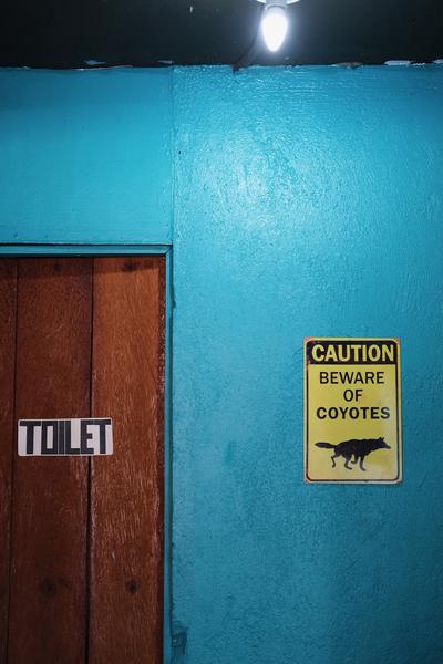 A door to a bathroom set in a blue wall. Next to the door is a sign reading “Caution. Beware of Coyotes.”