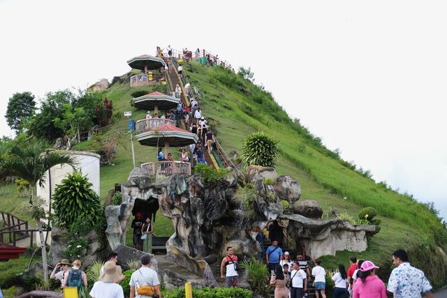 A view up a steep staircase cut into the side of a hill, leading to an observation deck at the top. Tourists pose for photographs at the base of the stairs. At regular intervals along the way there are shaded pavilions where visitors can rest.