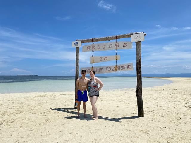 A woman and a man stand under a wooden sign reading “Welcome to Virgin Island”. A white sand beach stretches behind them to the horizon line.