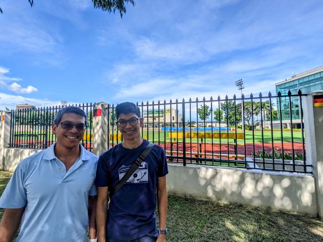 Two men in t-shirts smile, behind them is a concrete-and-iron fence that surrounds a high school athletic field