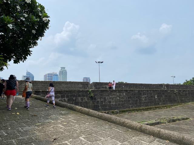 Tourists walk up a stone ramp leading to walls that surrounded Intramuros in Manila during the Spanish colonial era. In the distance the tops of skyscrapers peek over the stone walls.