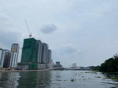 A view across a river at multiple high-rise buildings, one of which is covered by scaffolding and topped by a construction crane. The river is dotted with floating weeds.