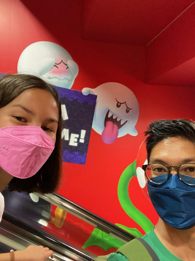 A father and daughter wearing masks pose for a selfie while riding an escalator. On the wall in the background are ghost characters from Nintendo’s Super Mario video games.