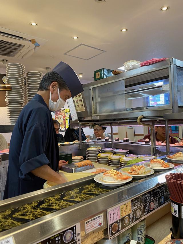 A chef preps sushi, surrounded by a conveyor belt of platters.