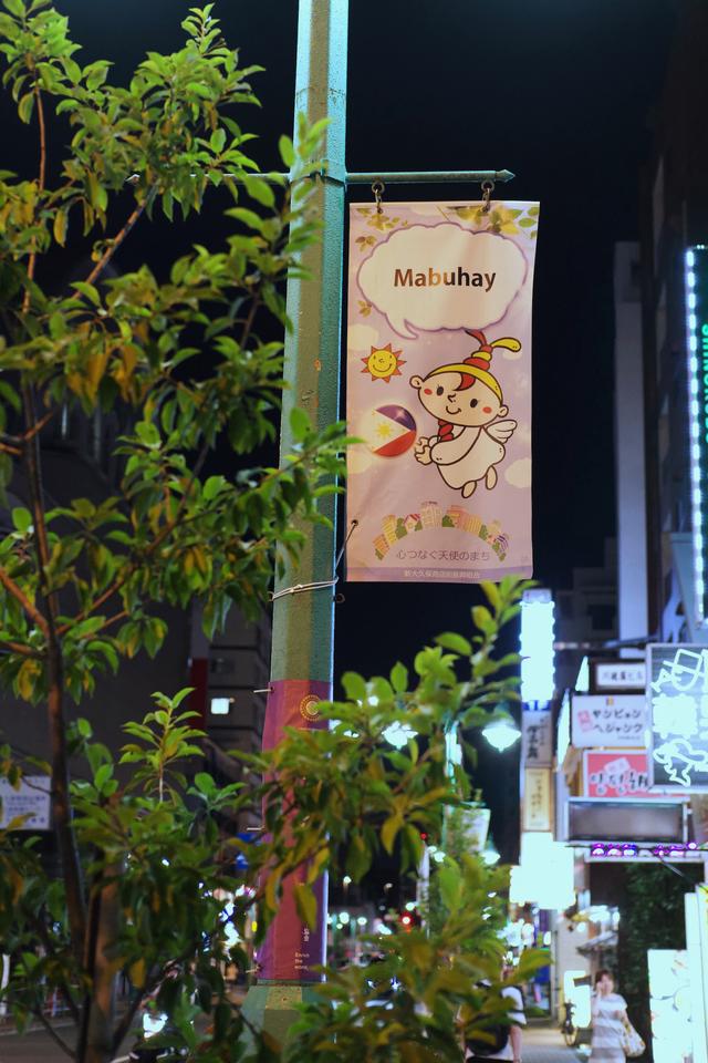 A banner hanging from a lamppost featuring a cartoon angel bearing a small Philippine flag. The angel has a speech bubble above it reading “Mabuhay”.