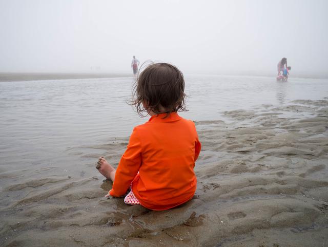 Young child in an orange shirt gazes out into fog while sitting on a beach.